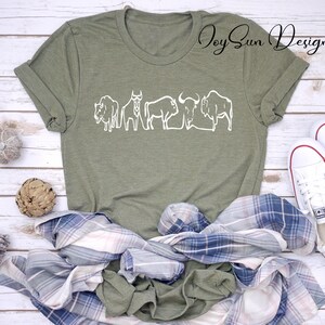 May include: Olive green t-shirt with a white line drawing of bison. The shirt is styled with a blue and white plaid shirt, and white sneakers. The text "Joy Sun Designs" is in the upper right corner.