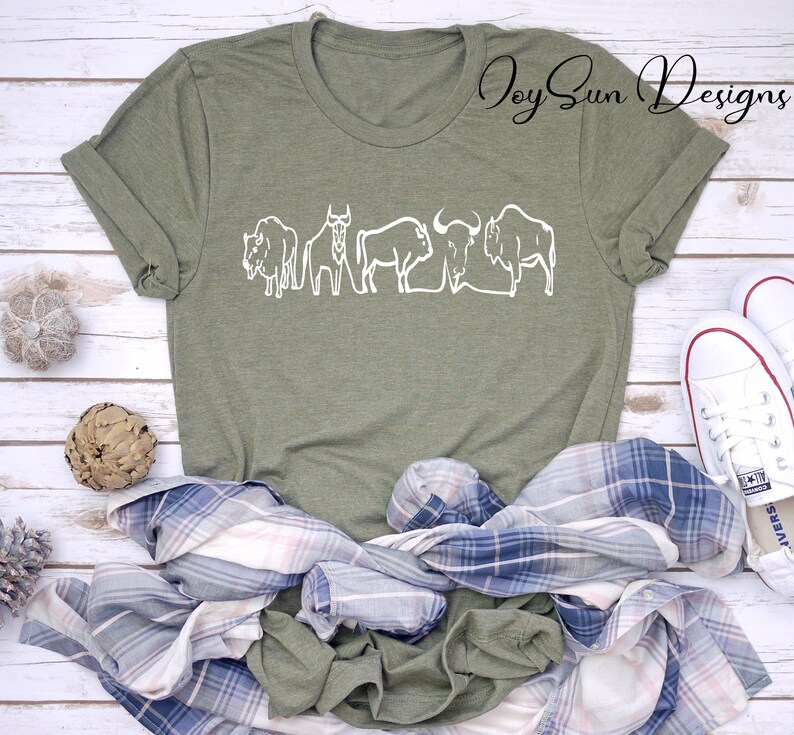 May include: Olive green t-shirt with a white line drawing of bison. The shirt is styled with a blue and white plaid shirt, and white sneakers. The text "Joy Sun Designs" is in the upper right corner.