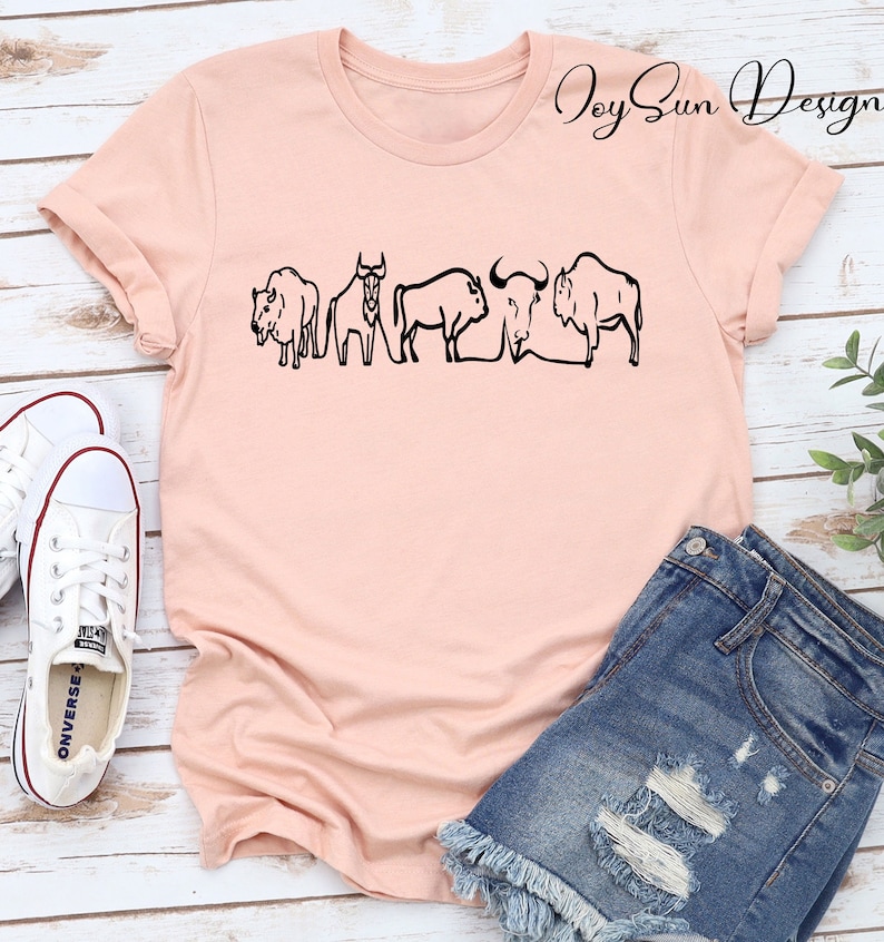 May include: A light pink t-shirt featuring a black line drawing of five bison. The shirt is paired with denim shorts and white canvas sneakers. The text "Joy Sun Design" is printed on the shirt.