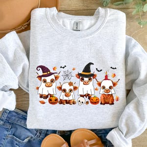 May include: Light gray sweatshirt featuring a Halloween-themed design. The graphic depicts four cartoon Highland cows dressed in costumes, including witch hats and ghost costumes, with pumpkins, bats, and a spiderweb. The sweatshirt is made of a soft material.