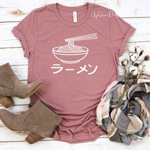 Puede incluir: Una camiseta rosa con un contorno blanco de un cuenco de fideos ramen y palillos. La palabra japonesa "Ramen" est&aacute; escrita en blanco debajo del cuenco.