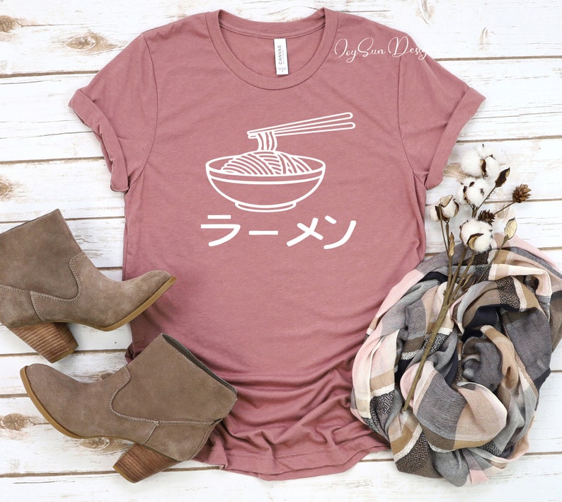 Puede incluir: Una camiseta rosa con un contorno blanco de un cuenco de fideos ramen y palillos. La palabra japonesa "Ramen" est&aacute; escrita en blanco debajo del cuenco.
