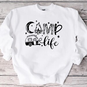 Pu&ograve; includere: Felpa bianca con testo nero che recita "Camp Life" con una grafica di un fal&ograve;, una luna e un camper.