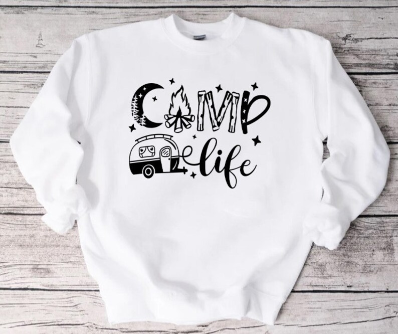 Pu&ograve; includere: Felpa bianca con testo nero che recita "Camp Life" con una grafica di un fal&ograve;, una luna e un camper.