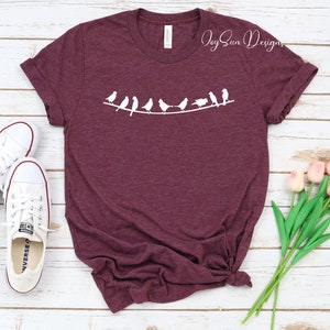 Peut inclure: Un t-shirt marron avec un motif blanc repr&eacute;sentant une ligne d'oiseaux sur un fil. Le texte "Joy Sun Designs" est visible dans le coin sup&eacute;rieur droit de l'image.