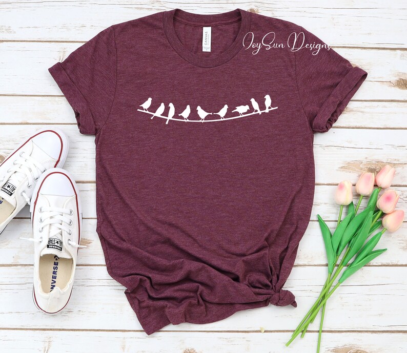 Peut inclure: Un t-shirt marron avec un motif blanc repr&eacute;sentant une ligne d'oiseaux sur un fil. Le texte "Joy Sun Designs" est visible dans le coin sup&eacute;rieur droit de l'image.