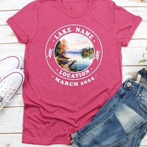 May include: A pink t-shirt with a white circle design featuring a watercolor illustration of a lake scene with a dock, trees, and mountains. The text "LAKE NAME", "LOCATION", and "MARCH 2024" is printed around the circle.