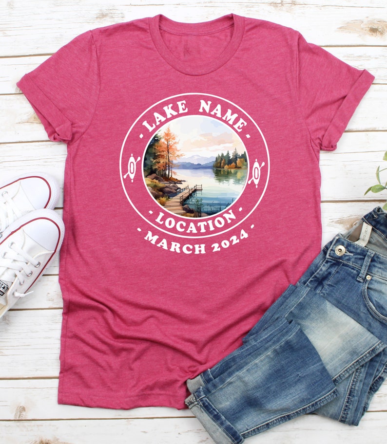 May include: A pink t-shirt with a white circle design featuring a watercolor illustration of a lake scene with a dock, trees, and mountains. The text "LAKE NAME", "LOCATION", and "MARCH 2024" is printed around the circle.
