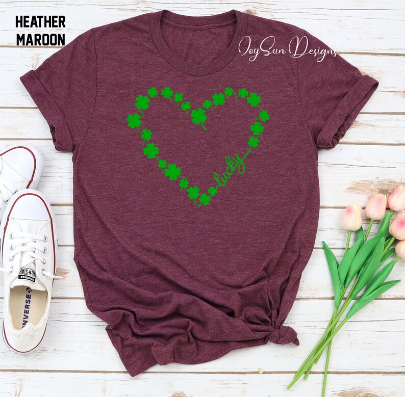 May include: Maroon t-shirt with a green heart made of shamrocks and the word "lucky". The shirt has rolled-up sleeves and is paired with white trainers. The text "Heather Maroon" is at the top left. The design is for St. Patrick's Day.