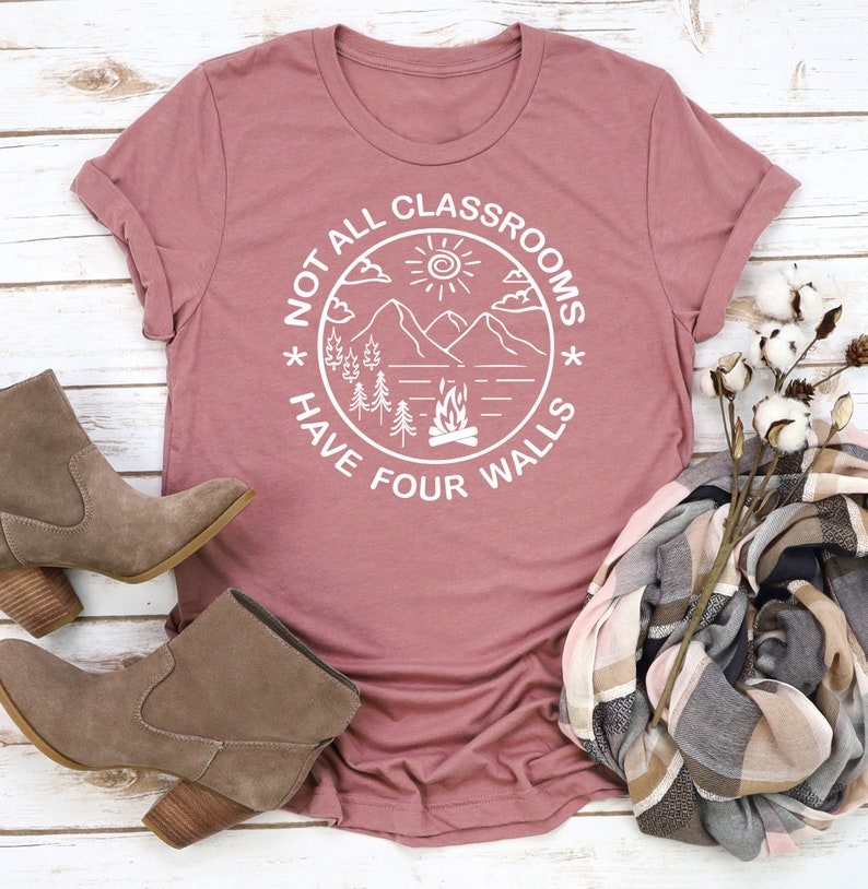 May include: A pink t-shirt with a white graphic design that says "Not all classrooms have four walls". The graphic features a mountain scene with a sun, clouds, trees, and a campfire.