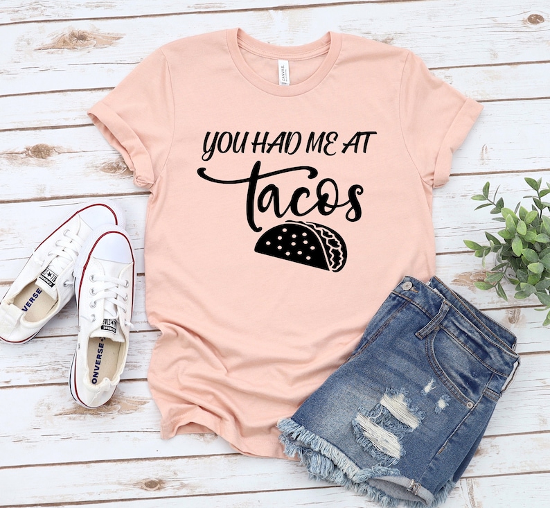 以下が含まれることがあります： ピンクのTシャツに黒い文字で「You had me at tacos」と書いてあり、黒いシルエットのタコスが描かれています。Tシャツは白い木製の表面に置かれ、白いスニーカーと青いデニムのショートパンツが置かれています。