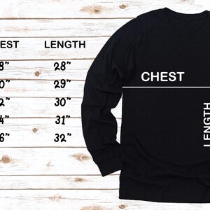 May include: Black long-sleeve shirt with size chart. The chart shows chest and length measurements in inches for sizes S to 2XL. The shirt has a crew neck and is displayed on a light wood grain background.