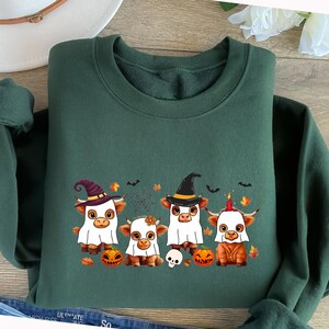 May include: A dark green sweatshirt featuring a Halloween-themed graphic. The graphic depicts four cartoon cows dressed in Halloween costumes, including a ghost, witch, and a cow with a candle on its head. Pumpkins, bats, and a spiderweb are also included.