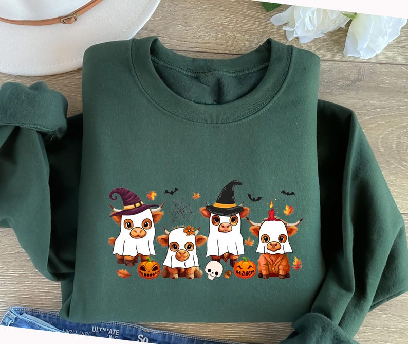 May include: A dark green sweatshirt featuring a Halloween-themed graphic. The graphic depicts four cartoon cows dressed in Halloween costumes, including a ghost, witch, and a cow with a candle on its head. Pumpkins, bats, and a spiderweb are also included.