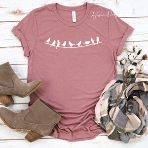 Peut inclure: Un t-shirt mauve avec un motif blanc de ligne d'oiseaux sur une branche. Le t-shirt est pos&eacute; &agrave; plat sur une surface en bois blanc avec une paire de bottines en daim marron et une &eacute;charpe &agrave; carreaux avec une tige de coton.
