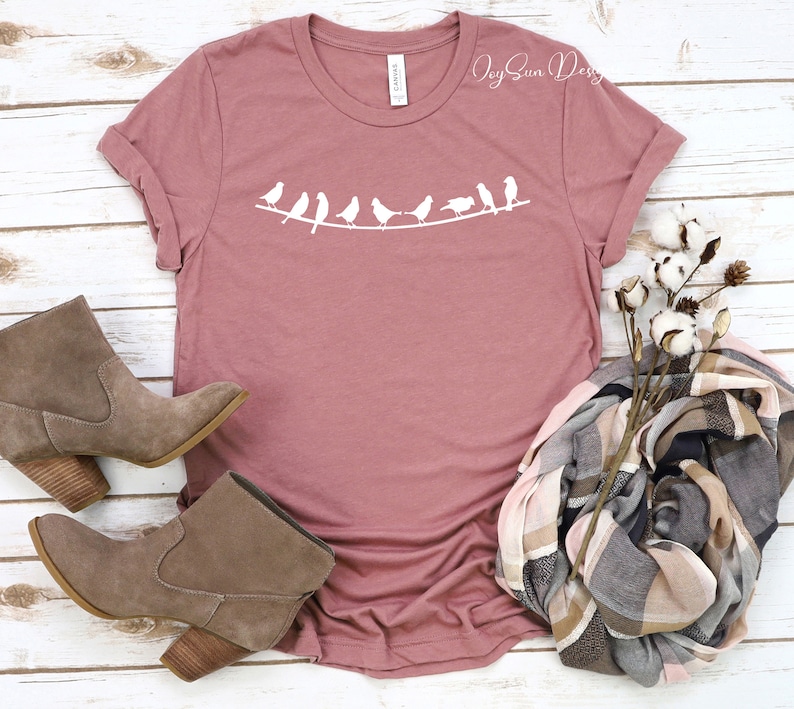 Peut inclure: Un t-shirt mauve avec un motif blanc de ligne d'oiseaux sur une branche. Le t-shirt est pos&eacute; &agrave; plat sur une surface en bois blanc avec une paire de bottines en daim marron et une &eacute;charpe &agrave; carreaux avec une tige de coton.
