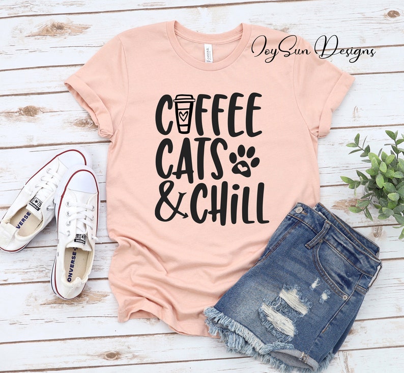 Puede incluir: Una camiseta rosa con el texto "Coffee Cats & Chill" en negro. El texto incluye una taza de caf&eacute; con un coraz&oacute;n y una huella de pata.