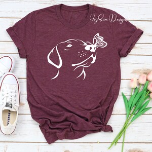 May include: A maroon t-shirt with a white silhouette of a dog's head with a butterfly on its nose. The text "Joy Sun Designs" is in the top right corner of the shirt.