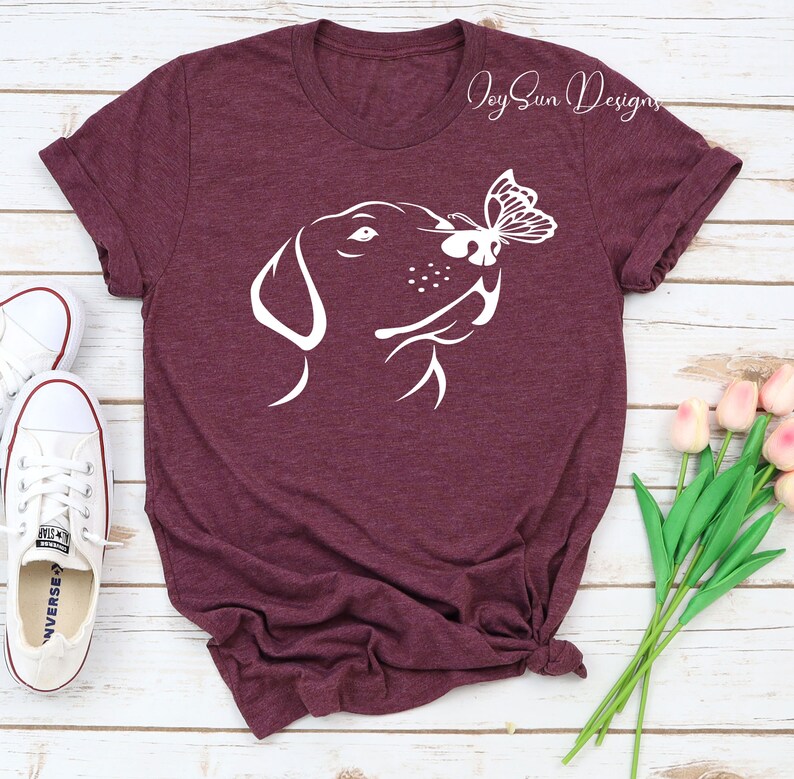 May include: A maroon t-shirt with a white silhouette of a dog's head with a butterfly on its nose. The text "Joy Sun Designs" is in the top right corner of the shirt.