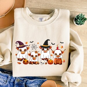 May include: Cream-colored sweatshirt featuring a Halloween-themed graphic. The design includes four cartoon cows dressed in costumes, including witch hats and ghost sheets. Pumpkins, bats, and a spiderweb are also part of the festive illustration.