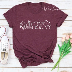 May include: Burgundy t-shirt featuring a white line drawing of several bison. The shirt has rolled-up sleeves and a knotted hem. White sneakers and pink tulips are visible in the background. The text "Joy Sun Designs" is in the upper right corner.