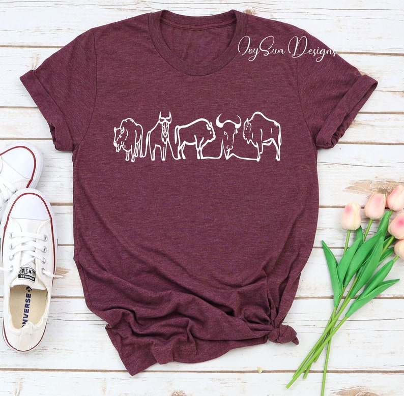 May include: Burgundy t-shirt featuring a white line drawing of several bison. The shirt has rolled-up sleeves and a knotted hem. White sneakers and pink tulips are visible in the background. The text "Joy Sun Designs" is in the upper right corner.