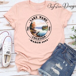May include: A pink t-shirt with a black circle design featuring a watercolor illustration of a lake scene with trees, a dock, and a canoe. The text around the circle reads "LAKE NAME", "LOCATION", and "MARCH 2024".