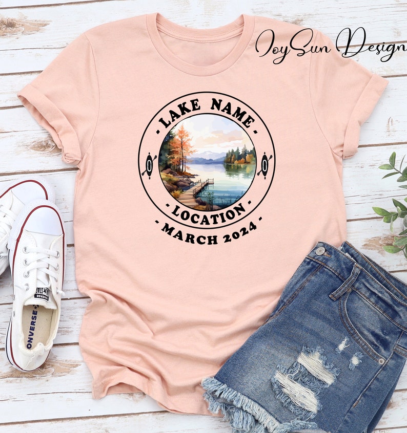 May include: A pink t-shirt with a black circle design featuring a watercolor illustration of a lake scene with trees, a dock, and a canoe. The text around the circle reads "LAKE NAME", "LOCATION", and "MARCH 2024".