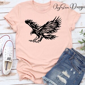 May include: A pink t-shirt with a black tribal style eagle design. The eagle is in flight with its wings spread wide. The design is centered on the shirt. The shirt is laying on a wooden surface with a pair of white sneakers and a pair of blue denim shorts.