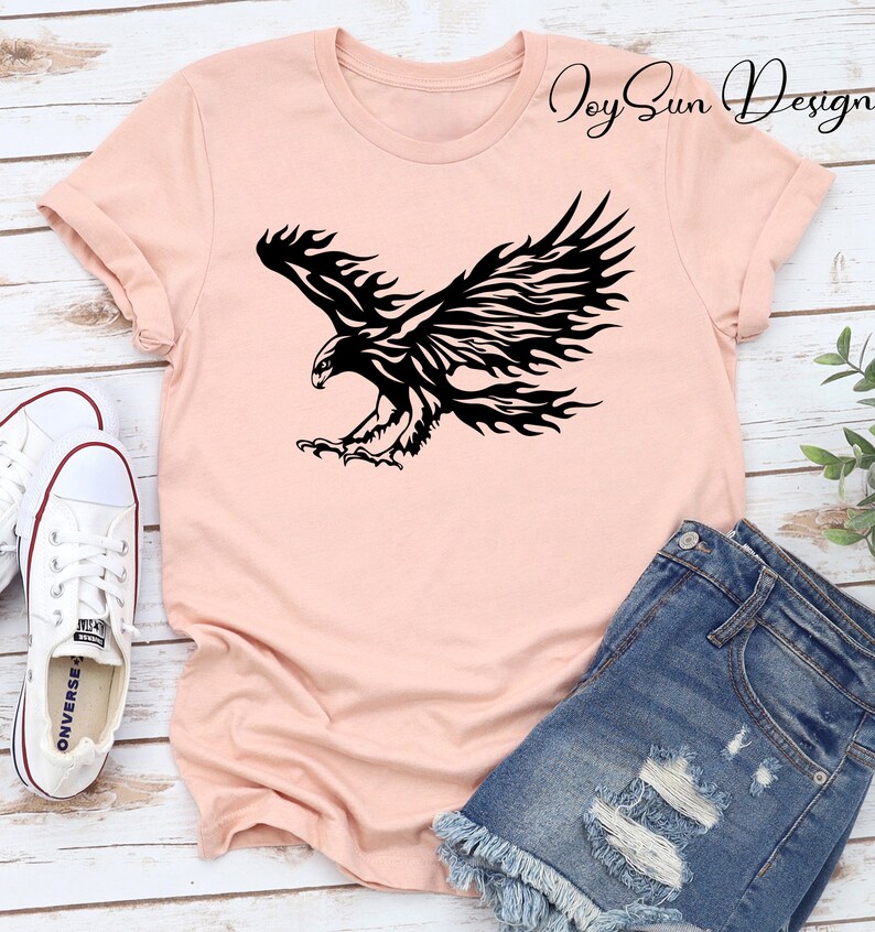 May include: A pink t-shirt with a black tribal style eagle design. The eagle is in flight with its wings spread wide. The design is centered on the shirt. The shirt is laying on a wooden surface with a pair of white sneakers and a pair of blue denim shorts.