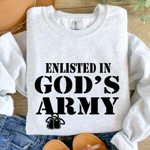 May include: A gray sweatshirt with the text "Enlisted in God's Army" in black lettering. The text is in a bold, blocky font. There are two small black dog tags hanging below the text.