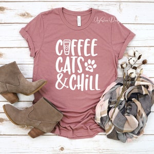 Puede incluir: Una camiseta color malva con el texto "Coffee Cats & Chill" en blanco. El texto incluye una taza de caf&eacute; y un gr&aacute;fico de huella de pata. La camiseta est&aacute; colocada sobre una superficie de madera blanca con un par de botines de ante marr&oacute;n y una bufanda de cuadros.