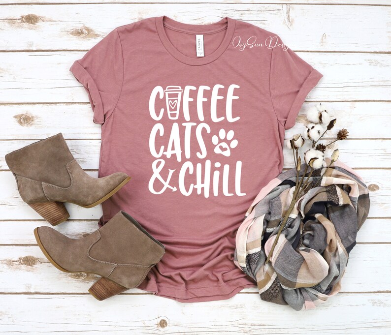 Puede incluir: Una camiseta color malva con el texto "Coffee Cats & Chill" en blanco. El texto incluye una taza de caf&eacute; y un gr&aacute;fico de huella de pata. La camiseta est&aacute; colocada sobre una superficie de madera blanca con un par de botines de ante marr&oacute;n y una bufanda de cuadros.