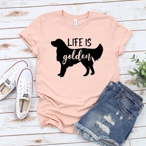 May include: A pink t-shirt with a black silhouette of a golden retriever and the text "Life is golden". The shirt is paired with denim shorts and white sneakers.