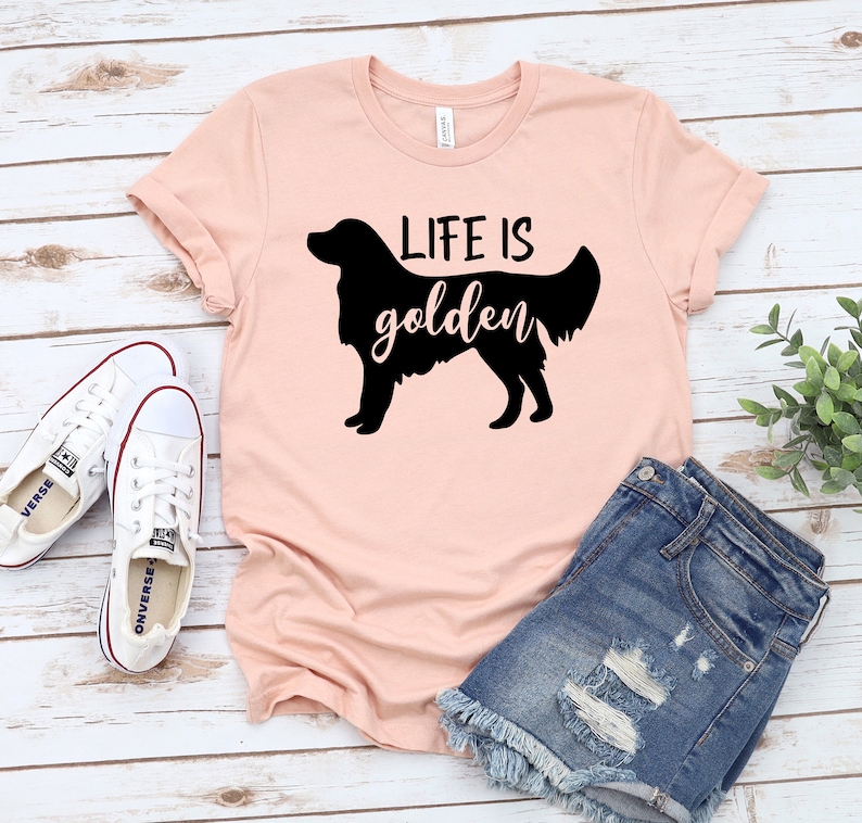 May include: A pink t-shirt with a black silhouette of a golden retriever and the text "Life is golden". The shirt is paired with denim shorts and white sneakers.