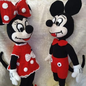 Op de afbeelding: Gehaakte Mickey Mouse en Minnie Mouse poppen. Mickey draagt een rode broek, een zwart shirt en gele schoenen. Minnie draagt een rode jurk met witte stippen, een bijpassende strik en gele schoenen. Beide poppen hebben zwarte oren en staarten.
