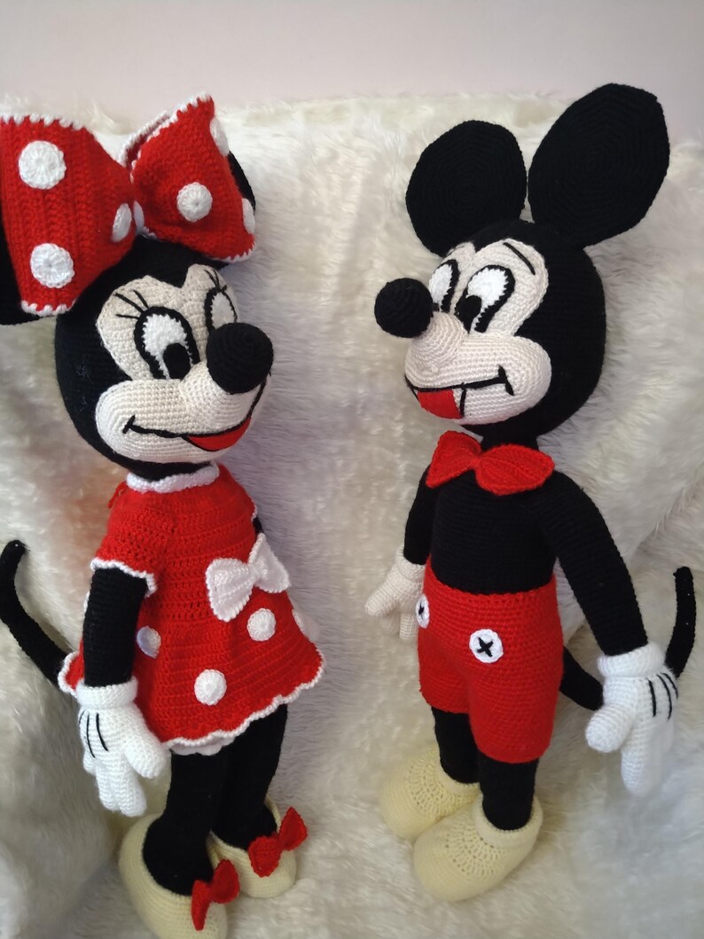 Op de afbeelding: Gehaakte Mickey Mouse en Minnie Mouse poppen. Mickey draagt een rode broek, een zwart shirt en gele schoenen. Minnie draagt een rode jurk met witte stippen, een bijpassende strik en gele schoenen. Beide poppen hebben zwarte oren en staarten.