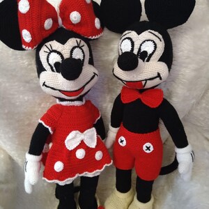 Op de afbeelding: Twee gehaakte poppen, Minnie en Mickey Mouse, staan naast elkaar. Minnie draagt een rode jurk met witte stippen en een bijpassende strik. Mickey draagt een rode korte broek, een zwart shirt en een rode vlinderdas. Beide hebben zwarte oren.