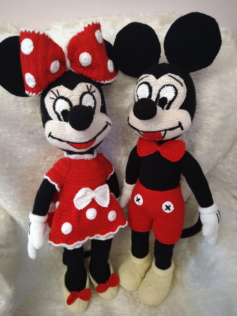 Op de afbeelding: Twee gehaakte poppen, Minnie en Mickey Mouse, staan naast elkaar. Minnie draagt een rode jurk met witte stippen en een bijpassende strik. Mickey draagt een rode korte broek, een zwart shirt en een rode vlinderdas. Beide hebben zwarte oren.
