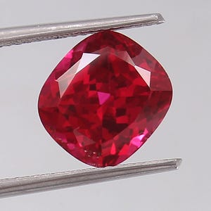 May include: A cushion-cut, faceted gemstone held by tweezers. The gem is a vibrant, deep red colour with multiple facets that reflect light. The shape is square with rounded corners, and the gem appears flawless.