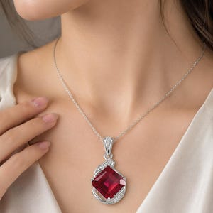 May include: A silver pendant necklace featuring a large, square-cut, deep red gemstone set in a polished silver frame. The setting is accented with small, clear, sparkling stones along the curved, asymmetrical silver edges of the pendant.