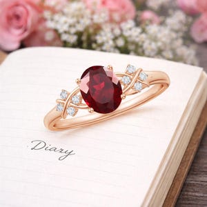 May include: A rose gold ring featuring a large, oval-cut deep red garnet gemstone at the centre. The band is accented on both sides by a cluster of four small, round, clear white diamonds arranged in a cross-like pattern, set in polished metal.
