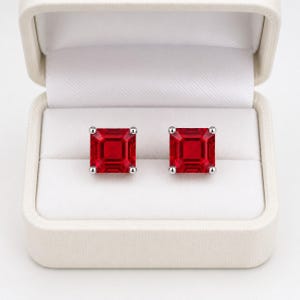 Könnte beinhalten: Ein Paar quadratisch geschliffene rote Edelstein-Ohrstecker, gefasst in silberfarbene Metallkrappen. Die Ohrringe sind nebeneinander in einer cremefarbenen Schmuckschatulle mit weißem Innenfutter und aufklappbarem Deckel präsentiert.