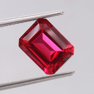 Peut inclure: Une pierre précieuse rectangulaire de taille émeraude d'une teinte rouge profonde et saturée. La pierre présente une grande clarté et des facettes géométriques nettes qui reflètent la lumière, maintenue par une pince métallique.