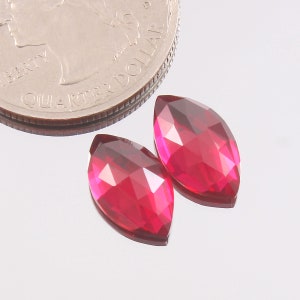 May include: Two faceted, marquise-cut, ruby-red gemstones next to a quarter coin. The gemstones have a high-gloss finish and reflect light, showcasing their multiple facets. The coin is silver and provides a size reference.
