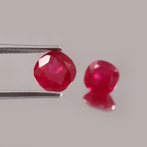 May include: Two faceted, deep red gemstones. One is held by silver tweezers, showcasing its brilliant cut. The other gem rests nearby, reflecting light. These rubies are a vibrant, rich color, perfect for jewelry.