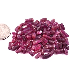 May include: A pile of raw ruby gemstones in varying shades of pink and red, with a quarter for size comparison. The rough-cut stones have a natural, unpolished appearance, showcasing their crystalline structure. The gemstones are scattered on a white surface.