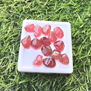 Puede incluir: Una colección de gemas en bruto, facetadas, de color rojo-rosa, expuestas sobre un plato cuadrado blanco. Las gemas tienen una cualidad translúcida y formas variadas. El plato se encuentra sobre un fondo de césped artificial verde.