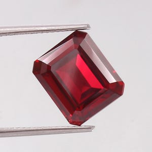 May include: A vibrant, deep red gemstone with a rectangular emerald cut, featuring sharp, clean facets that reflect light. The stone is held securely by a pair of silver-toned metal tweezers, showcasing its transparent clarity and rich crimson color.