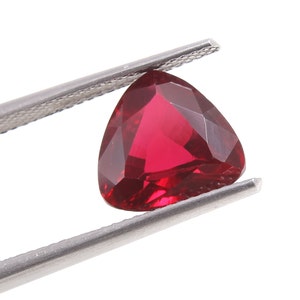 May include: A faceted, triangular-shaped, deep red gemstone is held by a pair of silver tweezers against a white background. The gem's cut reveals multiple facets, enhancing its brilliance and color.